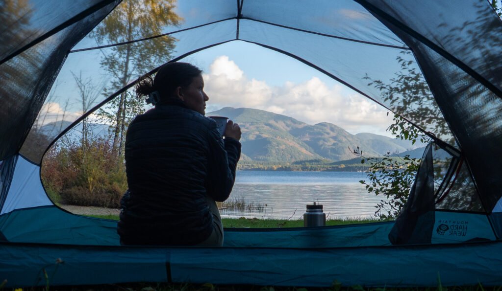 m Zelt mit Aussicht – Aus der Perspektive von innen: geöffnete Zelttür mit Blick auf Berge und See, mit frau die tee trinkt